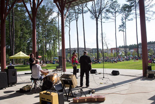 A band performs on an outdoor stage in Summerville, with musicians playing drums and guitars as a crowd watches from the grassy park area surrounded by tall pine trees and nearby buildings.